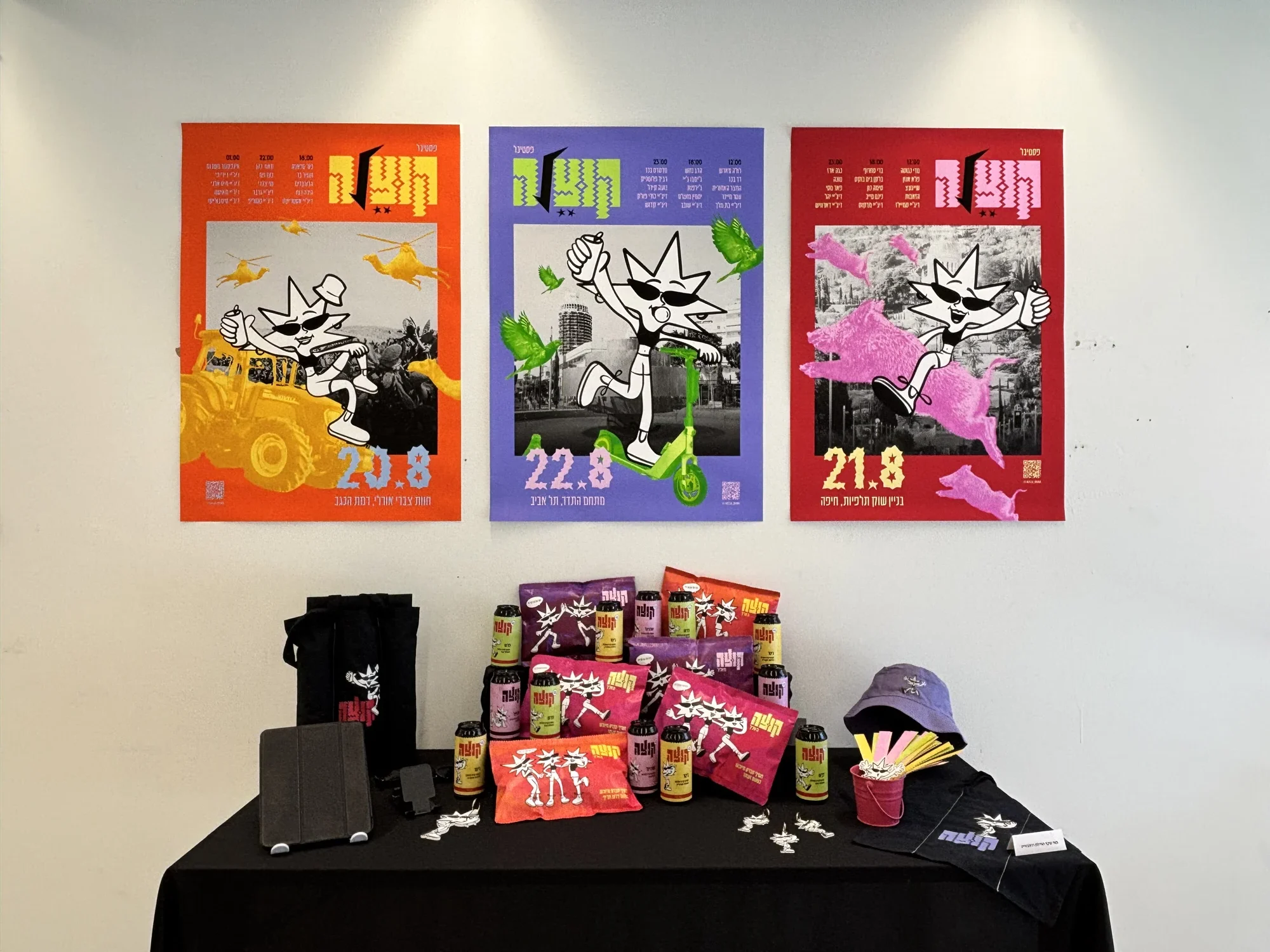 Top view of the Kutza launch table: cap, tote bags, cans and snack bags above three posters on a white wall